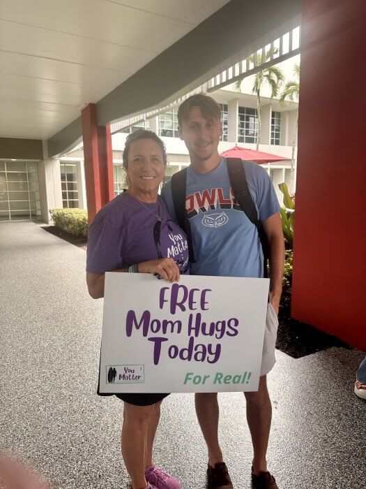 two people standing next to eachother lady holding sign that reads free mom hugs today